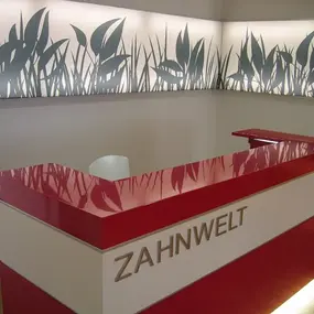Moderner Empfangstresen einer Zahnarztpraxis mit rotem Design und Naturdeko an der Wand.